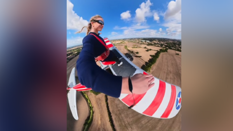 A blonde woman films herself wing-walking on a bi-plane with fields below.