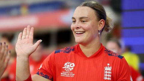 Danielle Gibson holds up her palm and is wearing a red England cricket shirt