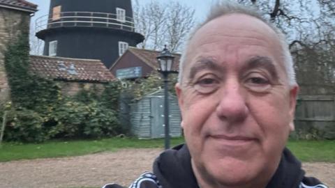 A man smiles as he takes a selfie in front of Alford windmill - a tall, black building surrounded by brick outbuildings and trees.