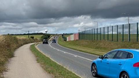 Several cars, including a blue hatchback, drive along a road on the boundary of a military base in Cornwall. The base has large fencing - including several sections coloured red and white - marking out the border. It is an overcast day.