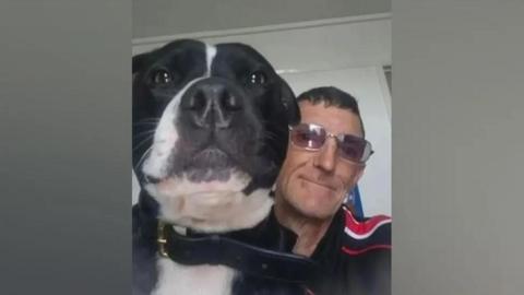 A dark-haired man wearing glasses and a black and red top, sitting with a black and white dog.