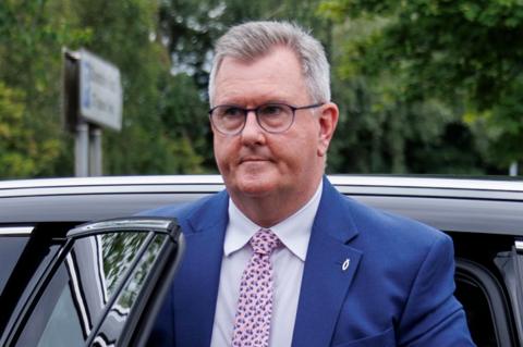 Jeffrey Donaldson is walking away from a black car. He is a man with short grey hair, wearing a dark blue suit, white shirt and pink tie.