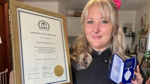 A woman with blond hair holding a gold-framed certificate and a silver medal in a blue presentation box. 