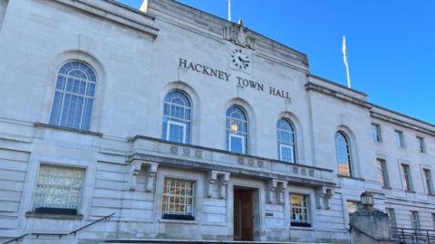 The art deco Hackney Town Hall building with Hackney Town Hall across the front under a clock face, with two rows of windows across the facade. 