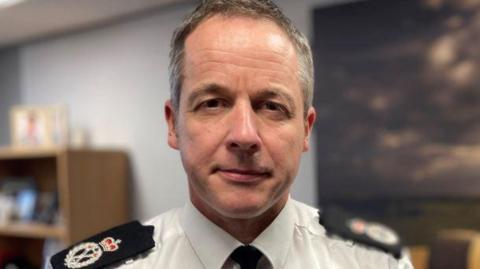 Chief Constable Paul Gibson. He has short receding grey hair and hazel-blue eyes. He is wearing a police uniform consisting of a white shirt with badges on the lapel and a black tie