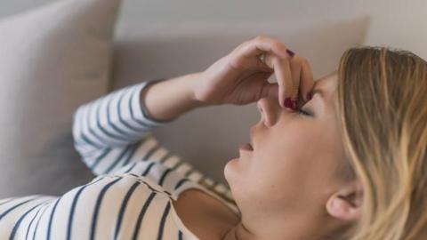 The image shows a young woman with blonde hair pinching the bridge of her nose with her eye closed, suggesting she has a headache or is suffering with some kind of fatigue. She is wearing a white top with black lines on it and she is laying on a grey sofa