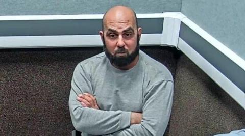 Amar Hussein is interviewed in a police station. He is largely bald with a trimmed black beard. He wears a light grey tracksuit and is sitting with his arms folded across his chest