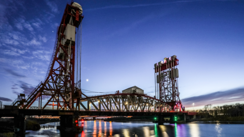 Newport vertical lift bridge - a metal structure consisting of two lifting pillars with the span of the bridge between them. The scene is early evening or late morning, with some light in the sky but the river reflecting the red and green lights on the base of each side of the bridge.
