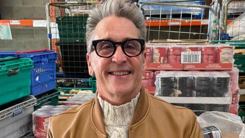 Neil Reid founder and chairman of The Big Food Project with grey hair wearing black rimmed glasses and a cream jumper and tan-coloured leather jacket in a warehouse of food