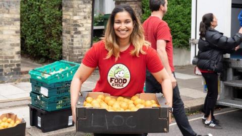 A lady, who has medium length brown hair, is wearing a red T-shirt which says FoodCycle on the front. She is holding a black box with potatoes in with both hands in front of her, and is standing on the side of a road.