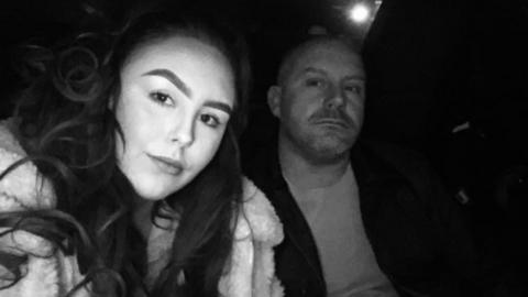 A black and white picture of Mollie Lord (left) and her dad Warren. She has long dark hair and wears a light coloured faux fur-style jacket. He has a bald head and wears a t-shirt and a black jacket