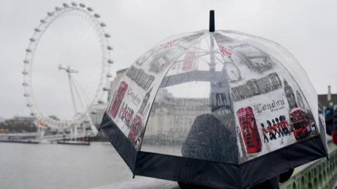 Someone stands with an umbrella with London symbols next to the Thames and the London Eye. It is raining.