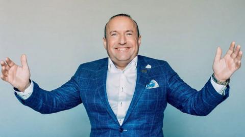 Dave Fishwick stands smiling widely with his arms outstretched to the sides/ He has very short dark hair and is wearing a blue checked suit jacket and a white shirt