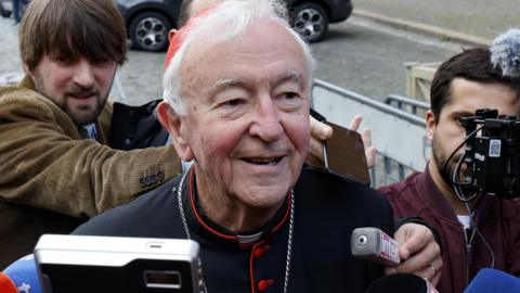 Cardinal Vincent Nichols has white hair and is wearing a red skull cap, black soutane with red trimmings, waistband and buttons as he is pursued by journalists on his way to a meeting with fellow cardinals in the Vatican.