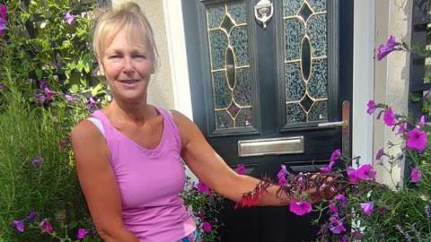 A woman with blonde hair and a full fringe tied back is wearing a bright pink vest and blue and pink shorts. She is smiling at the camera and standing next to a black and silver front door. Next to the front door are several purple flowers and greenery that she has planted.
