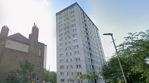 A white multi-storey building with a smaller stone building to the side. Cars are parked in the street outside