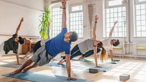 A man in a blue t-shirt doing pilates on an exercise mat surrounded by others doing the same.