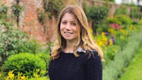 Rebecca Ableman smiling to the camera in a dark navy knitted jumper - there is a beautiful walled garden behind her