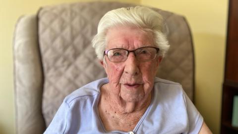 A woman with short white hair is looking at the camera. She is wearing rectangular metal glasses and a blue top. She is sitting in a chair.
