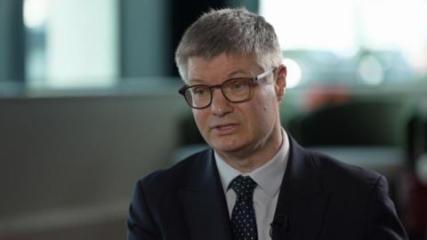 Lord Wolfson, chief executive of Next. He is sitting in a chair during an interview with the BBC. He is wearing glasses, a spotted tie, and a dark blue suit.