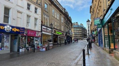 A row of shops in Halifax along a cobbled street. There are vape shops and discount stores to the left of the picture. On the right of the image there is a row of empty shops.