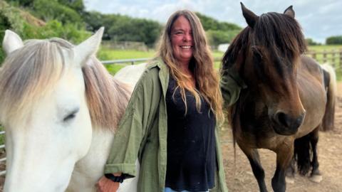 A woman with long hair smiles as she is flanked by a white horse and a brown horse 