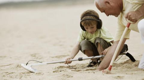 A young boy and an older man crouch in the sand, the boy holds a metal detector and then dig to see what is under the sand.