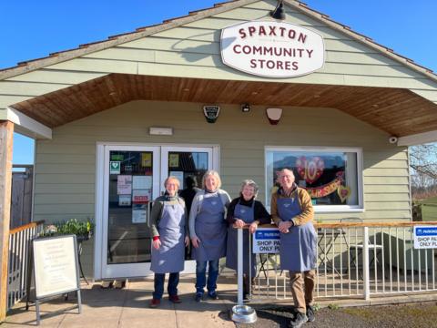 A group of four people in grey aprons stand outside a green building smiling together on a sunny day. 