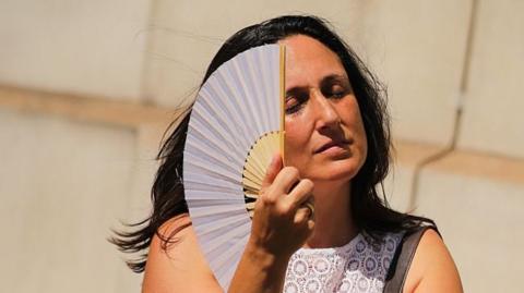 Woman has eyes closed as she fans herself in front of stone wall