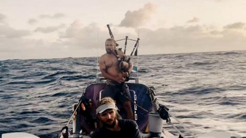A man in bare chest on a small boat in the sea plays the bagpipes.