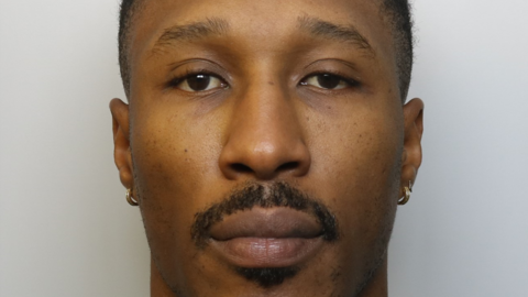 A man looking at the camera in a police mugshot, with a moustache and wearing two gold earrings.