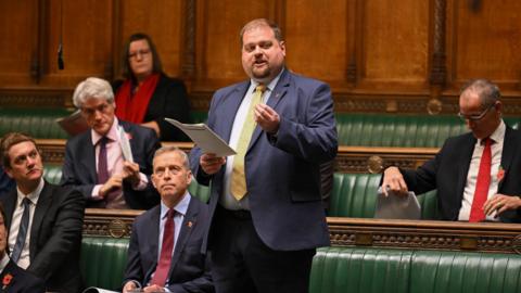 Dave Robertson, MP, stands up in parliament speaking, holding some papers. He has short hair and a beard, and is wearing a suit and a gold tie with a small poppy broach. There are green upholstered wooden benches behind him and four other men wearing suits, and a woman wearing a jacket and a scarf.