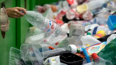 A stock image of a waste recycling centre with a bin filled with empty bottles and cartons. A woman can be seen adding a large Evian bottle to the pile.