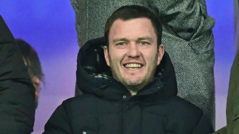 Craig Gardner sits in the directors' box in a dark puffer coat. He is smiling and behind him someone is standing up in a black and white tweed overcoat.