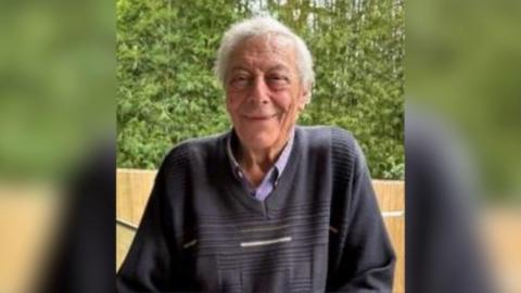 An elderly man with grey hair and wearing a dark blue jumper smiles at the camera