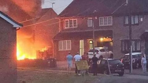 Several people stand outside in the street  looking towards a fire at a property.