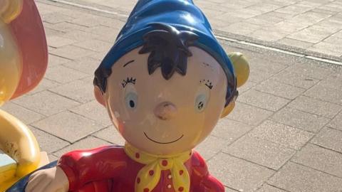 A plastic figure of Noddy, he has a blue hat on and red shirt with a yellow and red neckerchief. Noddy is positioned on a pavement, he is smiling at the camera. 