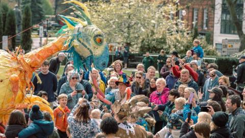 A large puppet of a phoenix made out of blue, yellow and orange material to the left of the frame. There is a crowd of people to the right smiling and looking at the puppet. They are in a grassy square with buildings on the right.