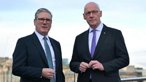 Sir Keir Starmer and John Swinney. Sir Keir has grey hair and Swinney has a bald head. They are both wearing glasses and dark suits with white shirts. Sir Keir is wearing a blue tie and Swinney a purple tie.