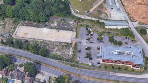 an aerial shot of a building, car park and unbuilt rectangle of land, next to a dual carriageway.