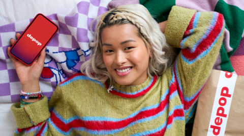 A posed photo of a young woman in a vibrant striped jumper, smiling for the camera with a phone showing the Depop logo