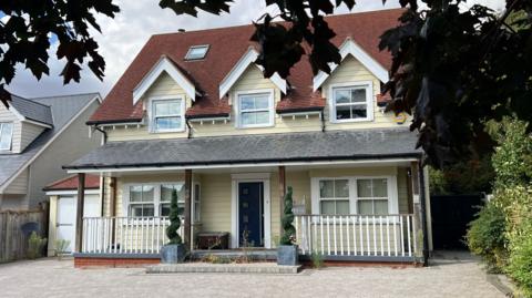 The front of a light yellow weatherboarded house of three storeys. It has a large driveway and two potted plants by the front door.