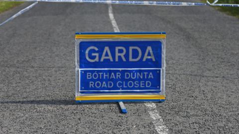 A blue sign placed on a road with white markings. The sign says Garda and road closed in both Irish and English.