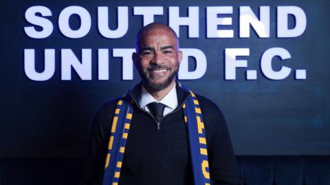 Kieron Dyer stands in front of a dark blue sign with white writing on it saying Southend United FC. He is wearing a zip-neck jumper with white shirt and dark tie underneath, with a blue and yellow scarf around his neck