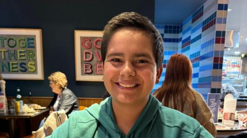 A boy smiles widely at the camera, he is wearing a green hoodie. He is sitting in a restaurant and other customers can be seen sitting on tables behind him