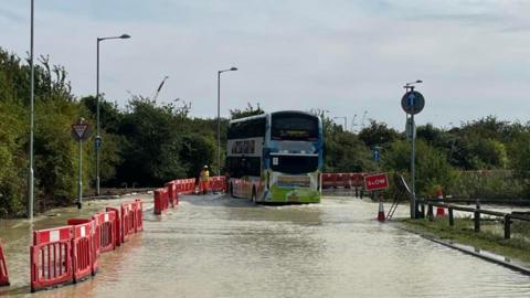 A double-decker bus is driving slowly through water which has flooded across a road. There are red barriers at the side of the road and a workman in a yellow jacket and hardhat can be seen in the distance close to the bus.