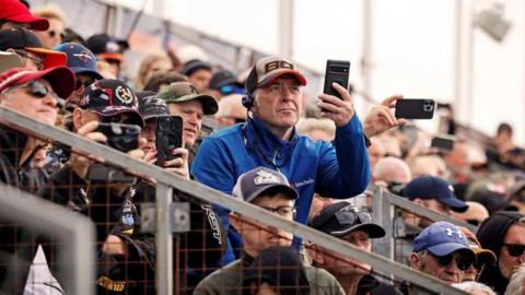 A crowd sitting on a grandstand, which sees men wearing TT caps. One wearing a bright blue jacket is standing up filming on his phone.