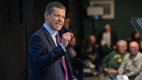 Rhun ap Iorwerth speaking at his party conference, in a dark suit, a blue shirt and a purple pokadot tie, in front of a teleprompter.