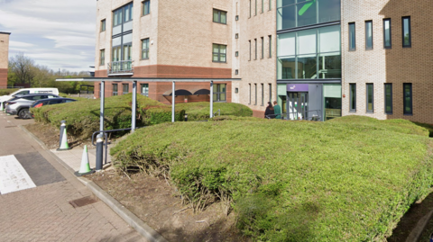 The office building is made of red and beige bricks. There is a green hedge and a zebra crossing out front.