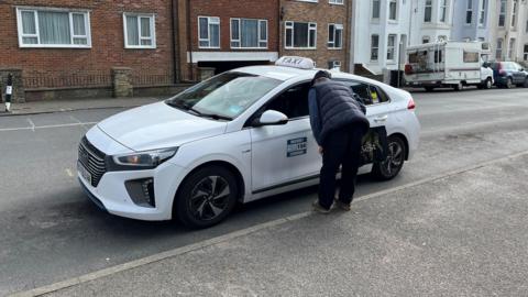 A white taxi waits at a rank in Seaford. An older man is speaking to the driver through the front passenger window.